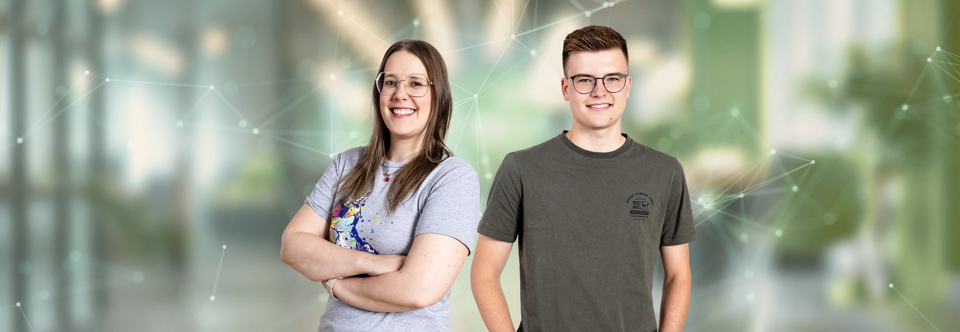 Eine Frau im hellgrauen T-Shirt mit verschränkten Armen und ein Mann im dunkelgrauen T-Shirt, beide mit Brille, lächeln in die Kamera. Dahinter liegt ein unscharfer, grünlicher Hintergrund mit feinen Netzwerklinien und Lichtpunkten.